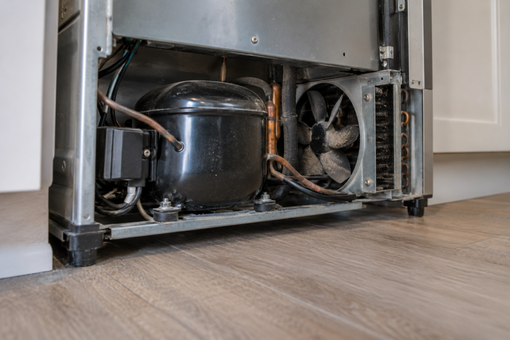 The compressor of an undercounter ice machine seen from floor level, with dust on the cooling fan blade beside it.